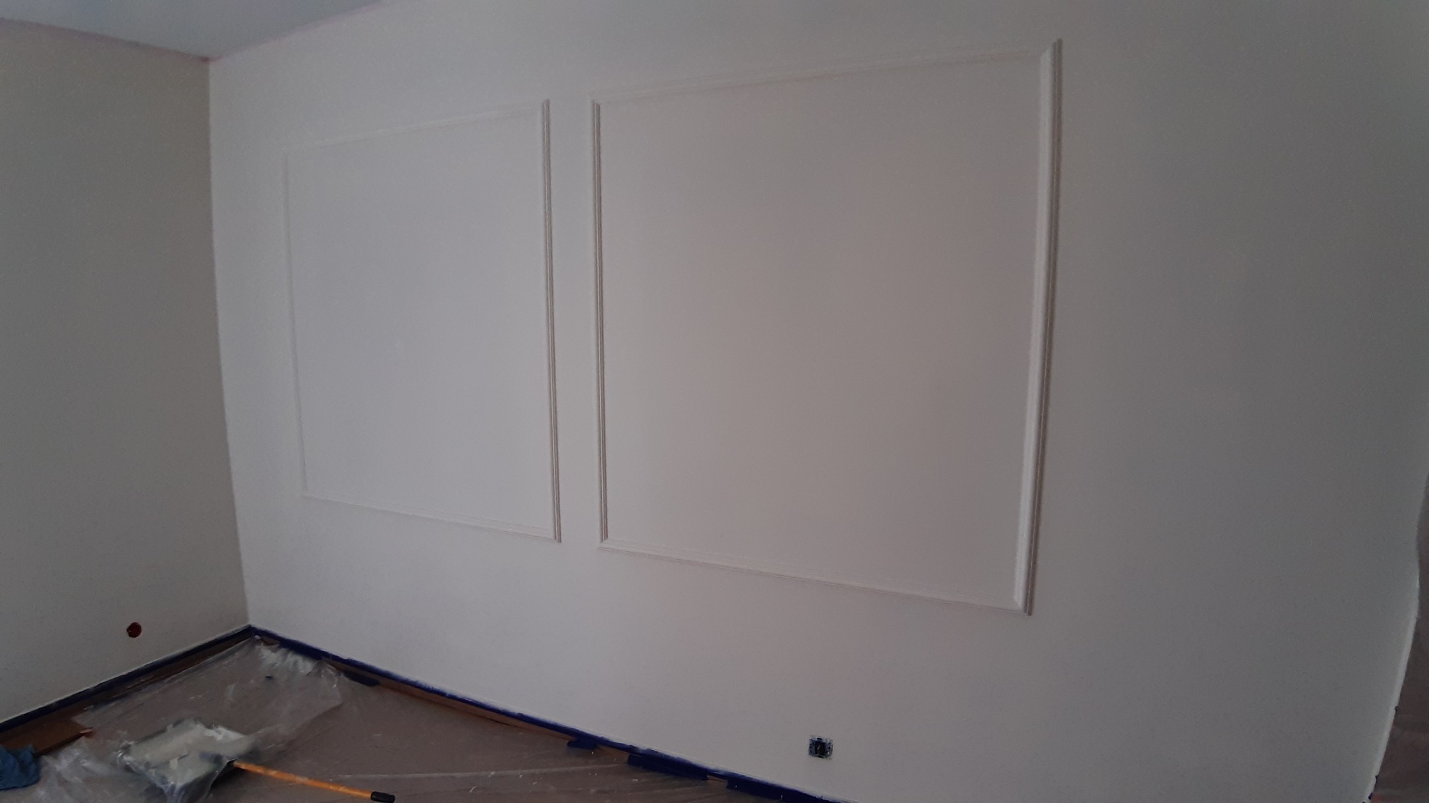 Świeżo pomalowana ściana z dekoracyjnymi ramkami, zabezpieczona folią podłoga, widoczny wałek malarski.