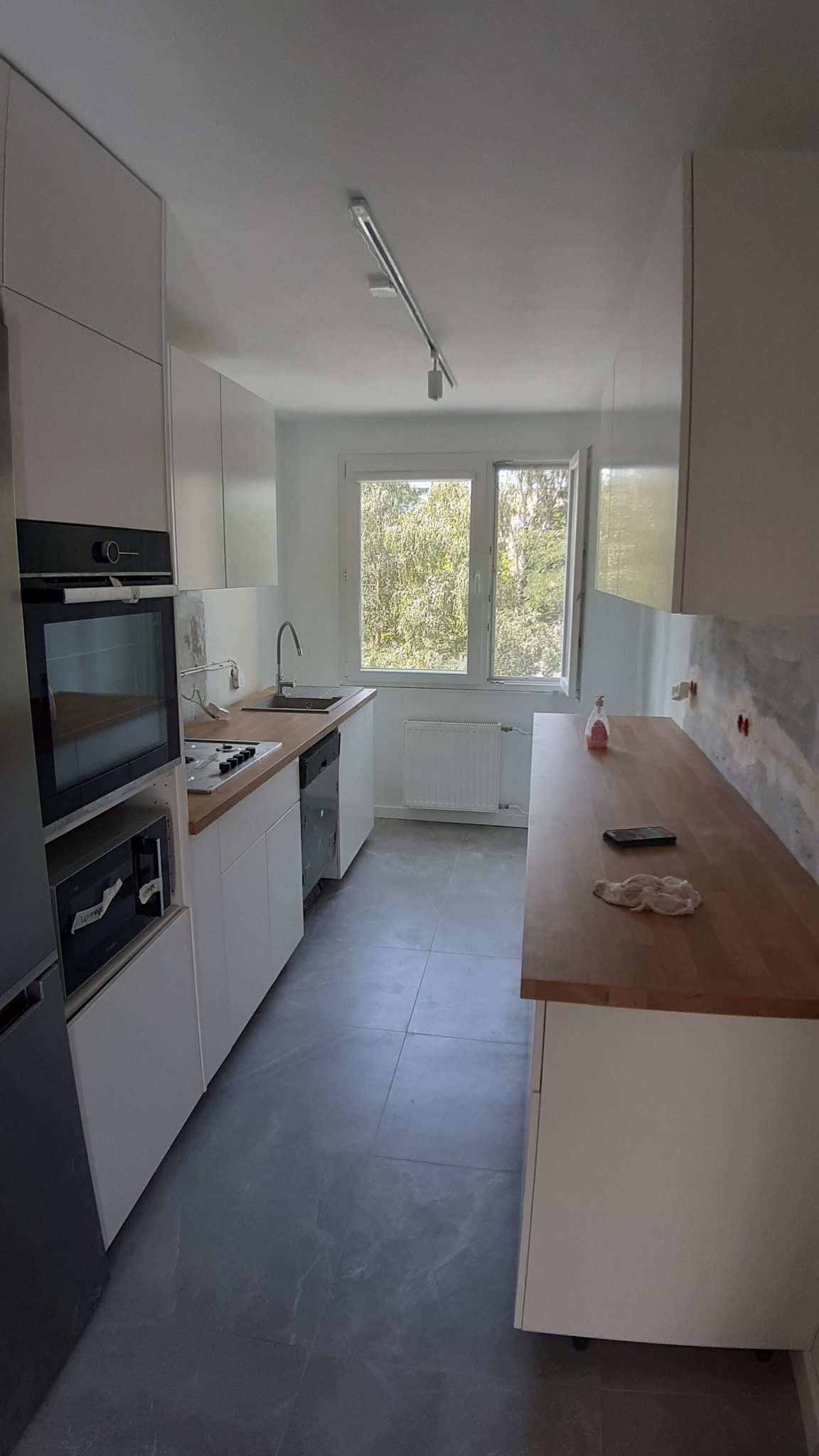 Nowoczesna kuchnia z białymi szafkami, drewnianym blatem i szarą podłogą z płytek imitujących marmur, widok na okno z zielenią na zewnątrz, ściana przygotowana pod płytki.