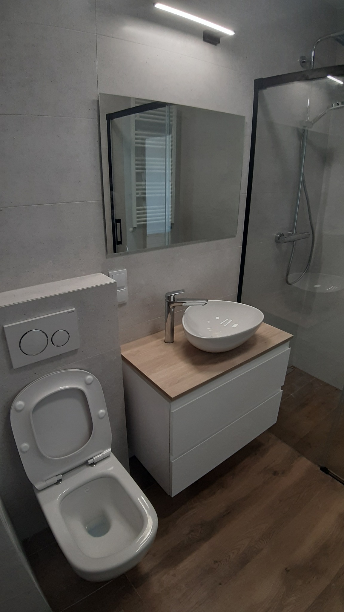 Nowoczesna łazienka z wiszącą miską WC, umywalką nablatową na szafce z drewnianym blatem i kabiną prysznicową z czarnymi ramami.