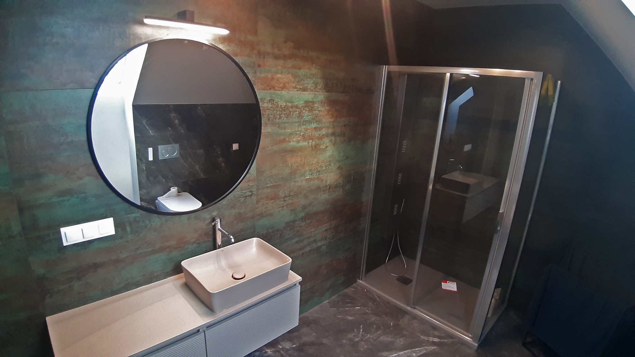 Nowoczesna łazienka z kabiną prysznicową, okrągłym lustrem, umywalką nablatową i płytkami imitującymi rdzę na ścianie.