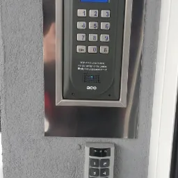 Zewnętrzny panel domofonowy ACO z klawiaturą numeryczną w srebrnej ramce, zamontowany na szarej, chropowatej ścianie obok drugiego, mniejszego panelu Safet.