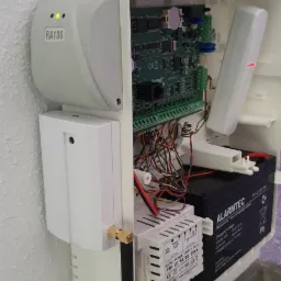 Otwarta obudowa centrali alarmowej RA100 z widoczną płytą główną, akumulatorem Alarmtec i zasilaczem, zamontowana na ścianie z listwą przypodłogową.