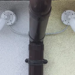 Dwie białe kamery monitoringu marki Novus zamontowane symetrycznie po obu stronach brązowej rury spustowej na elewacji budynku o nierównej fakturze.