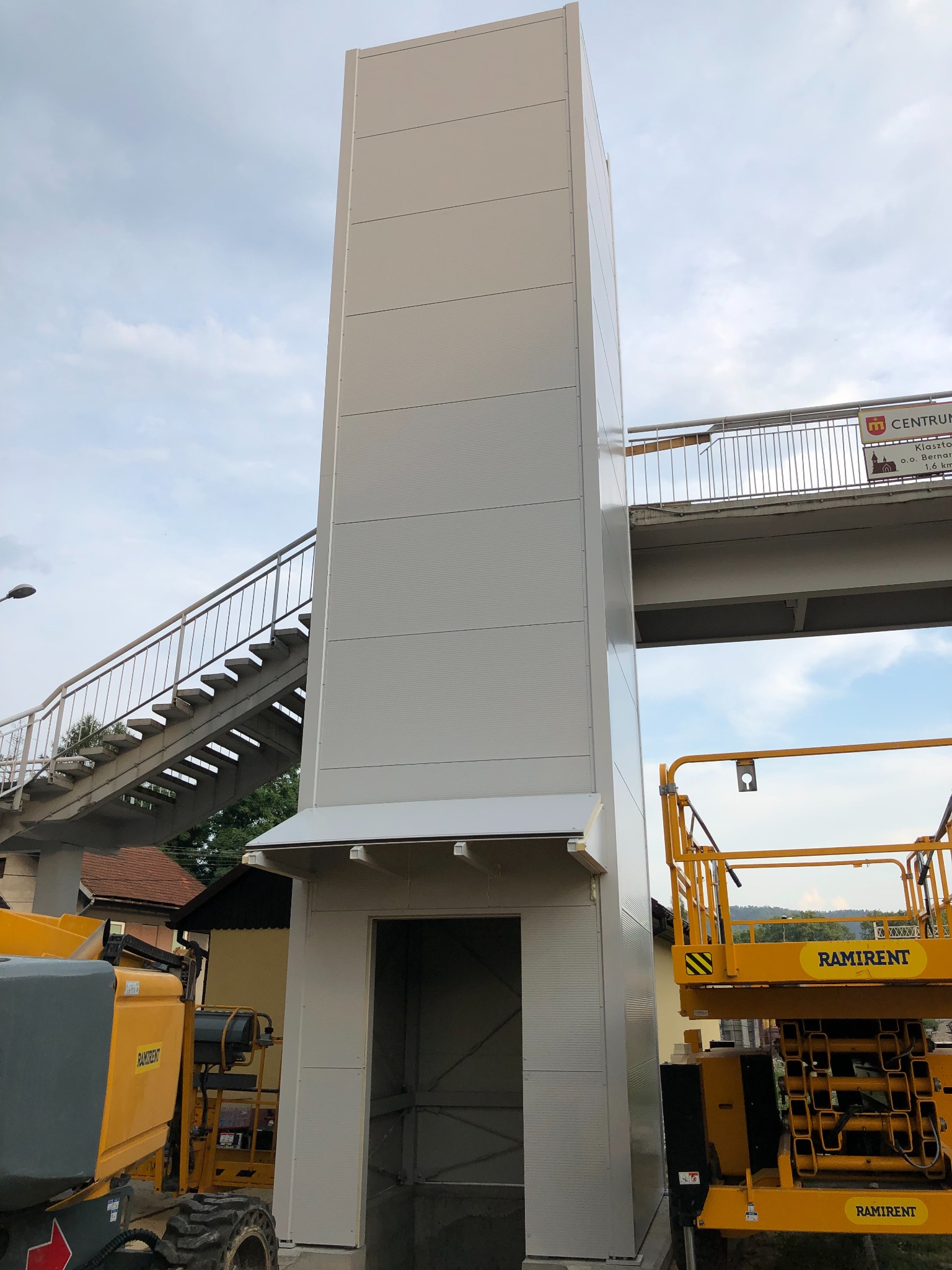 Zewnętrzna winda dla pieszych, obudowana panelami warstwowymi, z widocznymi podnośnikami koszowymi Ramirent w trakcie montażu przy kładce dla pieszych.