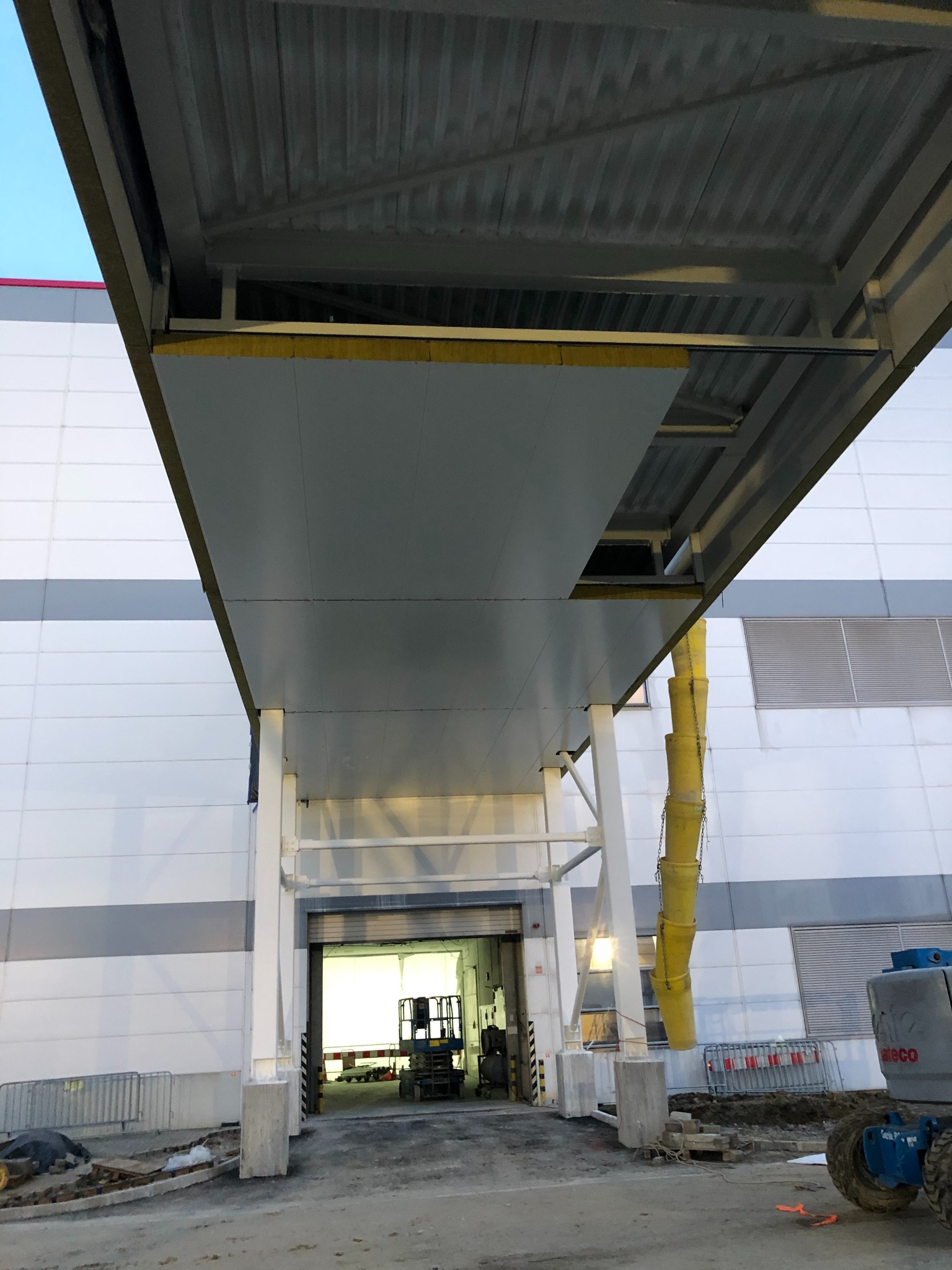 Zadaszone przejście nad bramą w hali produkcyjnej, widok od dołu, widoczne płyty warstwowe, konstrukcja stalowa, suwnica wewnątrz hali, zsyp budowlany i podnośnik koszowy na zewnątrz.