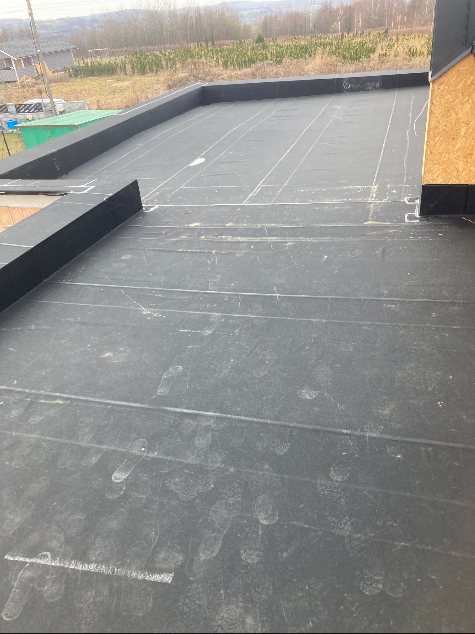 Wykończony, płaski dach pokryty czarną membraną z widocznymi śladami stóp. W tle drzewa i budynki. Elementy wykończeniowe z blachy i płyty OSB.