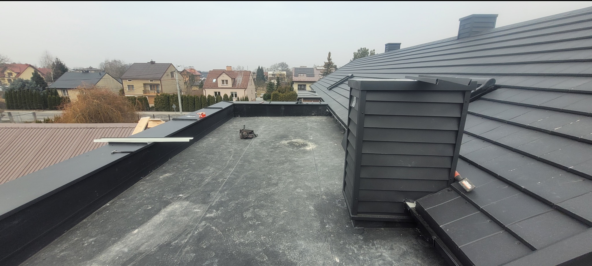 Czarny dach z blachy na budynku jednorodzinnym, widok z góry. Widoczny komin obłożony deskami, narzędzia dekarskie i zabudowa dachu płaskiego. W tle zabudowa domów.