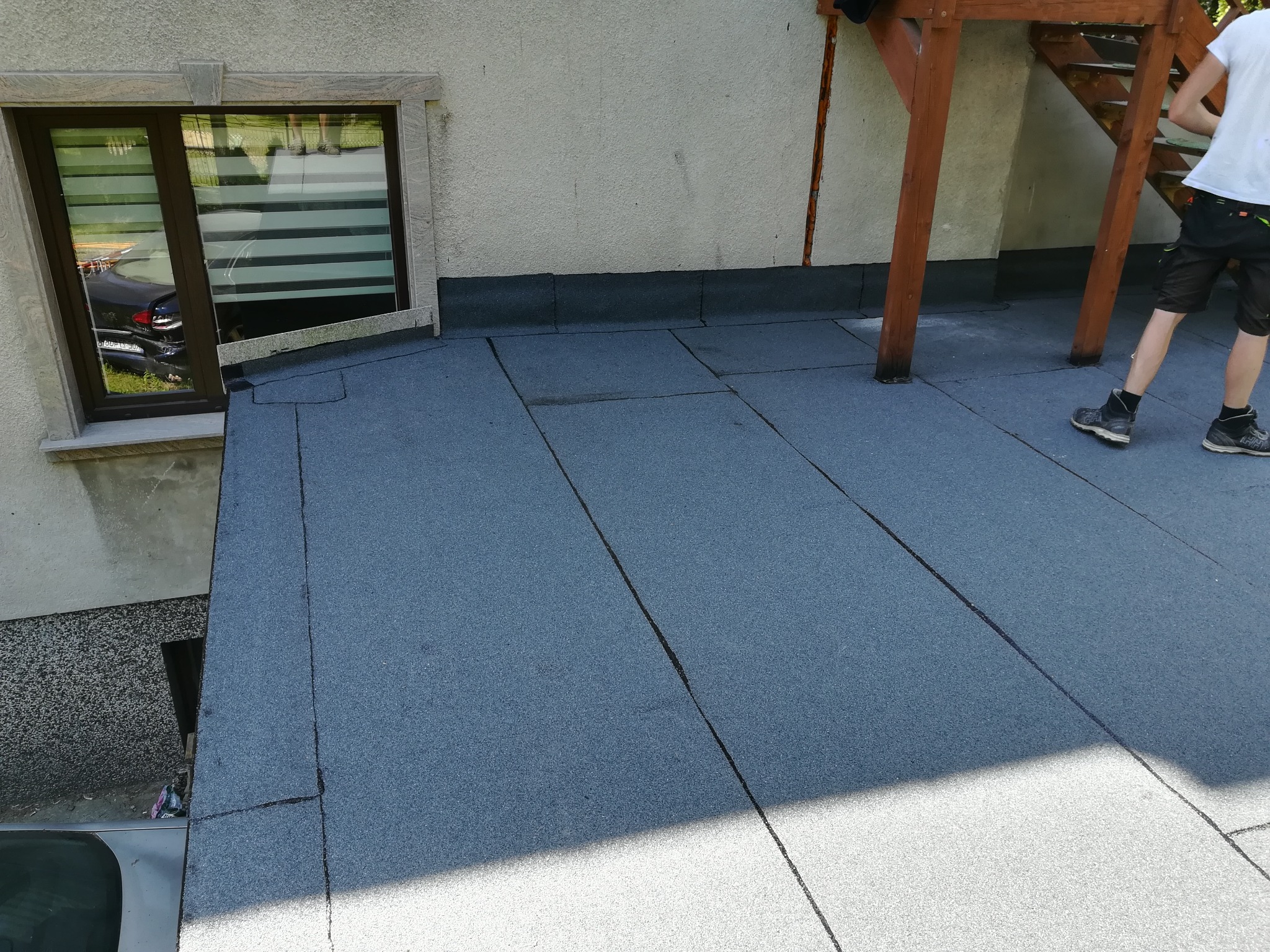 Szary, płaski dach pokryty papą termozgrzewalną, widok z góry. Fragment okna i schodów zewnętrznych. Osoba w tle. Jasne oświetlenie.