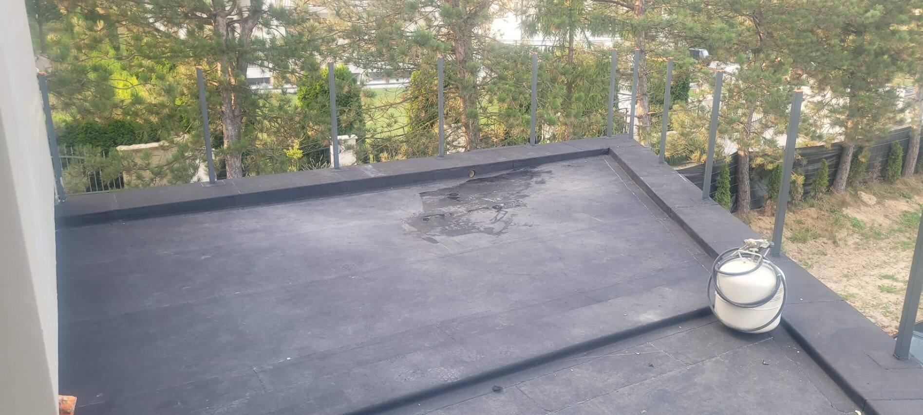 Czarny, płaski dach pokryty papą z widocznymi śladami wody, zabezpieczony metalową barierką. Butla gazowa leży na dachu. W tle zieleń drzew i krzewów.