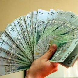 Dłoń trzymająca wachlarz z banknot&oacute;w 100 PLN na rozmytym tle, widoczne numery seryjne i znaki wodne na banknotach.