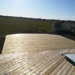 Nowo wybudowany drewniany taras z widokiem na zielone pole i szklarnię w słoneczny dzień. Część tarasu ułożona pod kątem, obok paczka materiałów budowlanych.