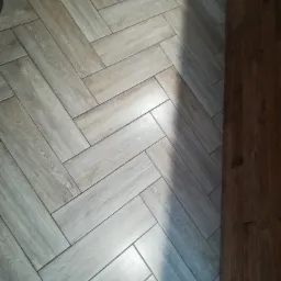 Ułożona w jodełkę podłoga z jasnych, drewnopodobnych płytek ceramicznych, z widocznym fragmentem ciemniejszej, drewnianej podłogi przy krawędzi, oświetlona naturalnym światłem tworzącym cień.