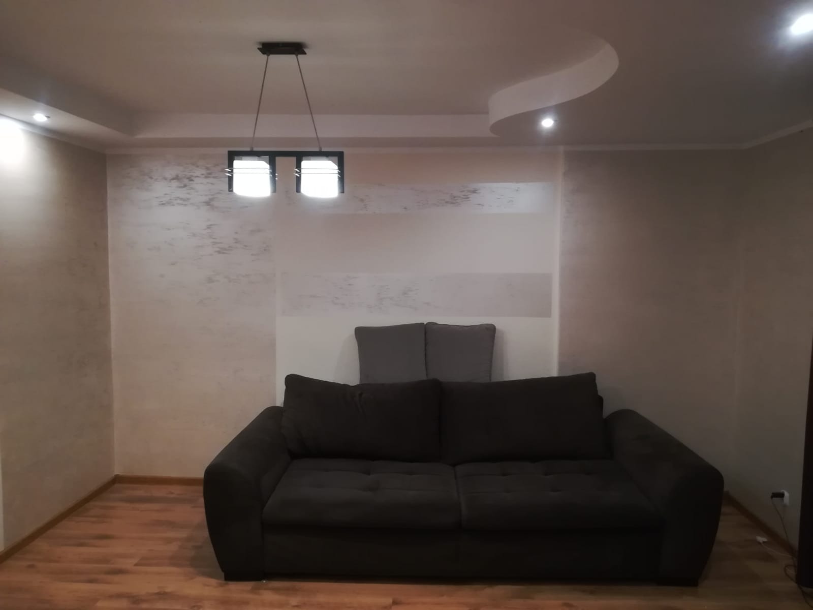 Pokój dzienny z ciemną sofą na drewnianej podłodze, ściany z dekoracyjną strukturą, nowoczesna lampa wisząca i dekoracyjny element sufitowy z oświetleniem punktowym.