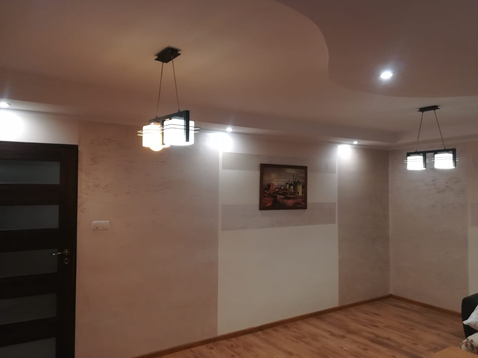 Jasne wnętrze pokoju z drewnianą podłogą, ścianami z dekoracyjną fakturą w odcieniach beżu i bieli, obrazem w ramie oraz dwiema nowoczesnymi lampami sufitowymi z białymi kloszami, widoczne gładkie...