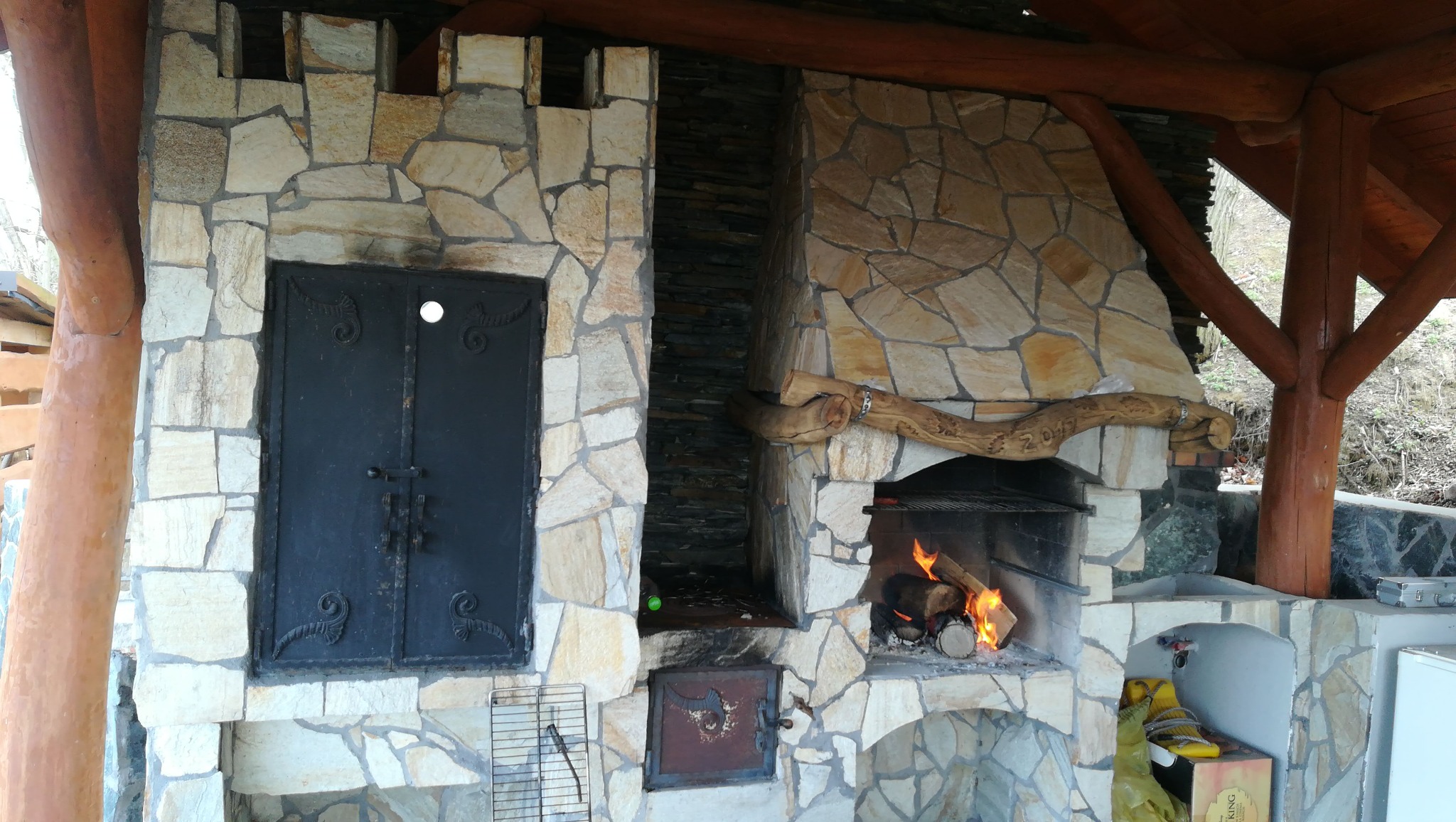 Kamienny grill ogrodowy z czarnymi, zdobionymi drzwiczkami, paleniskiem z płomieniami i drewnianą konstrukcją zadaszenia.