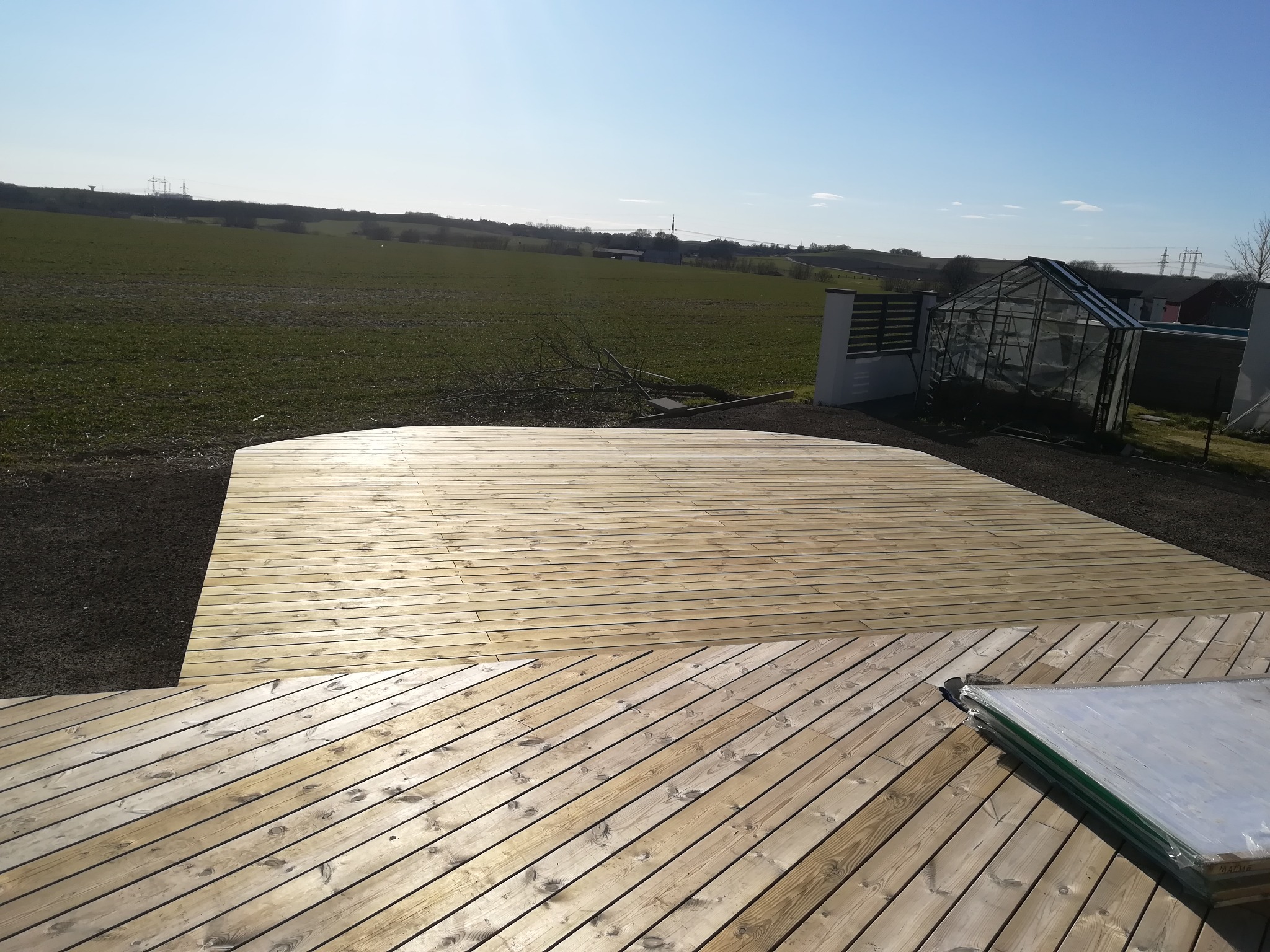 Nowo wybudowany drewniany taras z widokiem na zielone pole i szklarnię w słoneczny dzień. Część tarasu ułożona pod kątem, obok paczka materiałów budowlanych.