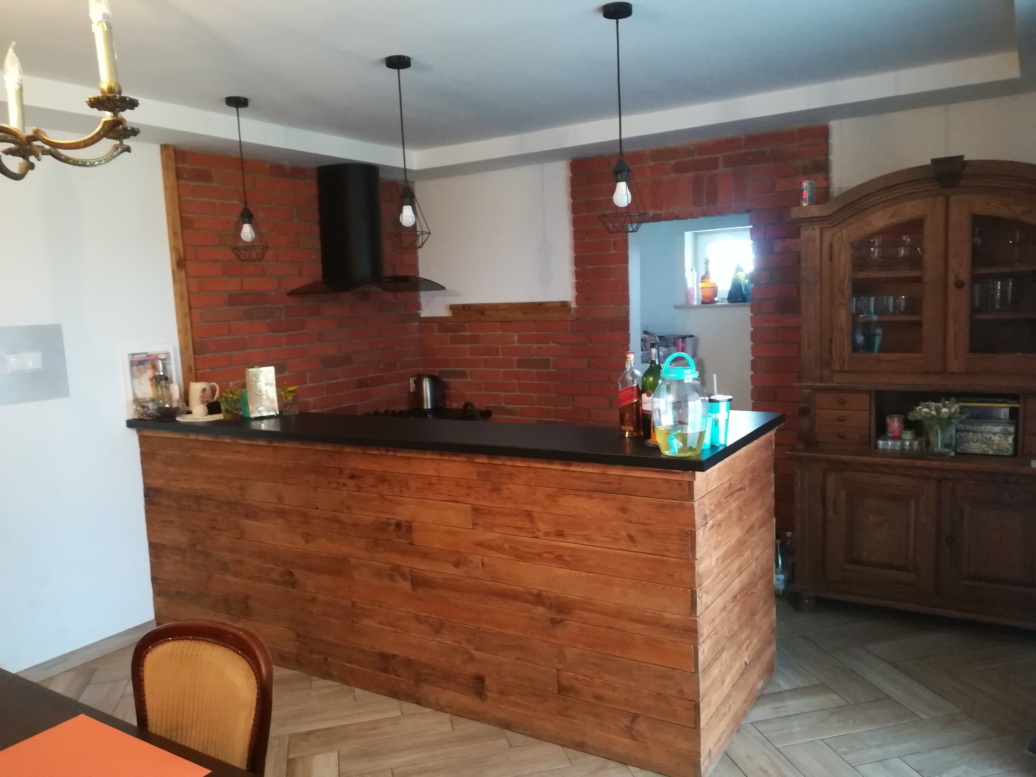 Kuchnia z ceglaną ścianą, drewnianym barkiem z czarnym blatem, trzema wiszącymi lampami i starodawną szafką z przeszklonymi drzwiczkami, widok na fragment stołu z pomarańczową podkładką.