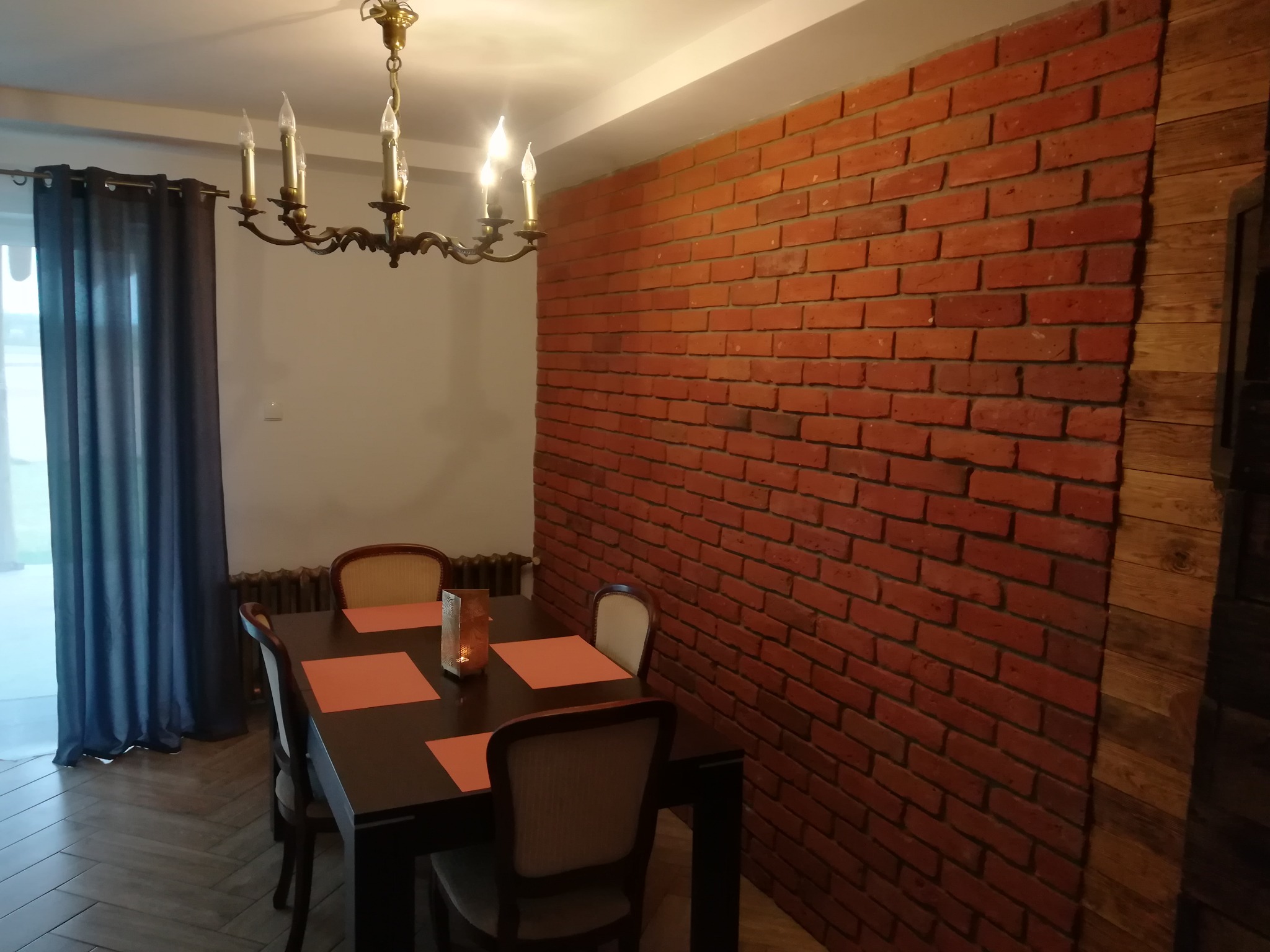Ceglana ściana w jadalni z drewnianym panelem, oświetlona świecznikiem, stół z krzesłami i zasłony w tle.