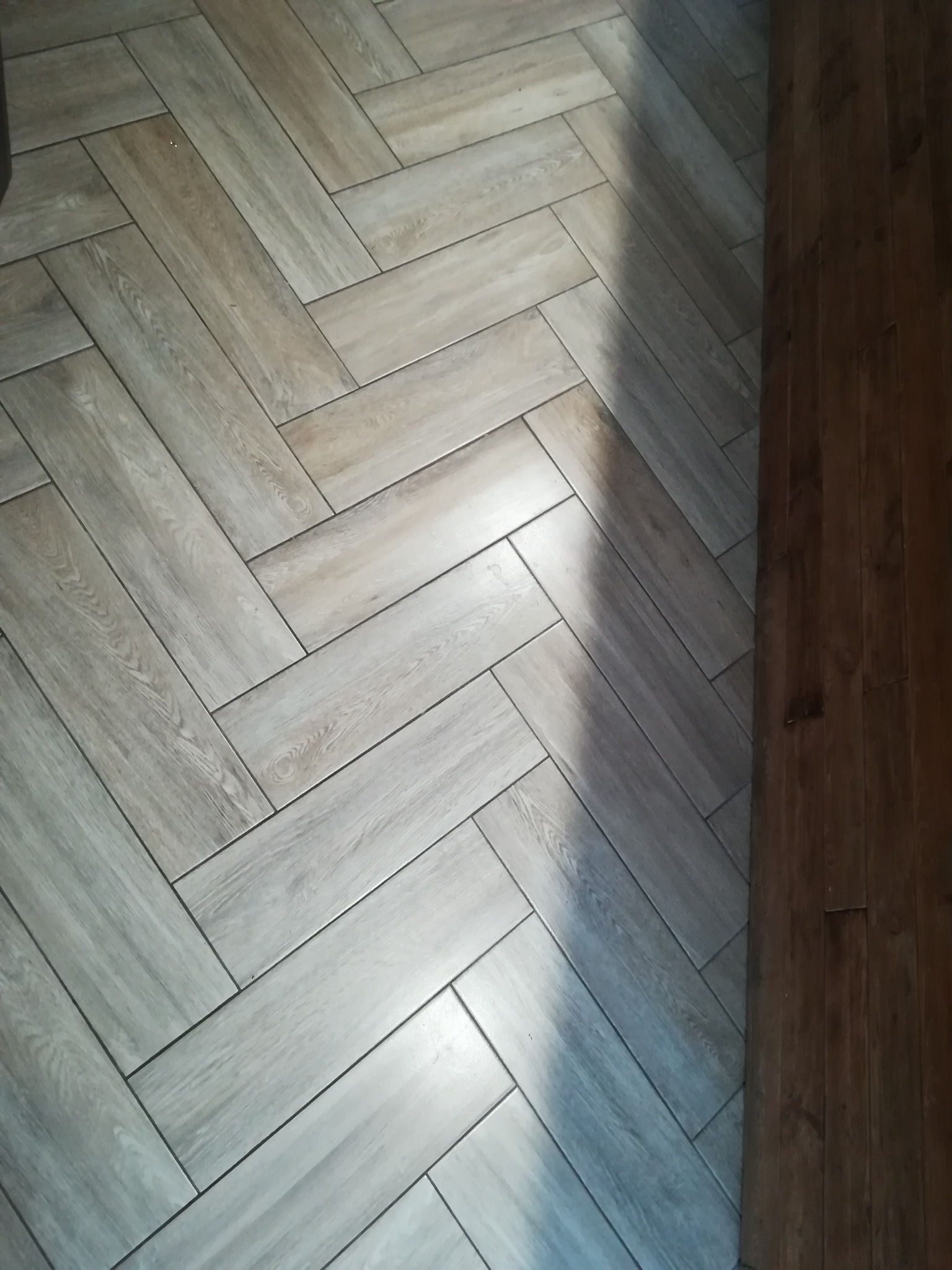 Ułożona w jodełkę podłoga z jasnych, drewnopodobnych płytek ceramicznych, z widocznym fragmentem ciemniejszej, drewnianej podłogi przy krawędzi, oświetlona naturalnym światłem tworzącym cień.