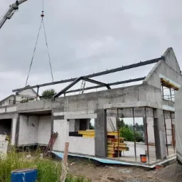 Montaż stalowej konstrukcji dachu na surowym budynku z betonu komórkowego, widoczny dźwig i materiały budowlane na placu budowy.