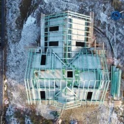 Widok z góry na budowę domu w stanie surowym otwartym, z widoczną konstrukcją ścian i dachu, częściowo pokrytą szronem, obok widoczny ogród i sąsiedni dom.