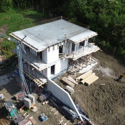 PM-BAU Budownictwo Energooszczędne - Wysoka perspektywa na budowę domu w stanie surowym otwartym z żurawiem, materiałami budowlanymi i częściowo wylanym fundamentem. Otoczenie to drzewa i ziemia.