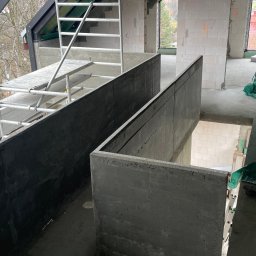 PM-BAU Budownictwo Energooszczędne - Wnętrze domu w trakcie budowy: surowe ściany z betonu komórkowego, rusztowanie, palety z bloczkami H+H. Widok z góry na betonowe elementy konstrukcyjne.