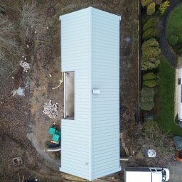 PM-BAU Budownictwo Energooszczędne - Nietypowy widok z drona na dom w trakcie budowy z szarą elewacją i fragmentem dachu. Widoczny niezabudowany otwór okienny i biały samochód dostawczy.