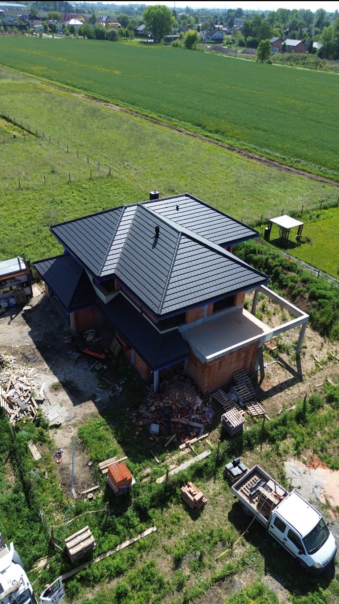 Dom w trakcie budowy z ciemnym dachem z blachodachówki, widok z drona. Częściowo otynkowane ściany z czerwonej cegły, elementy konstrukcji betonowej.