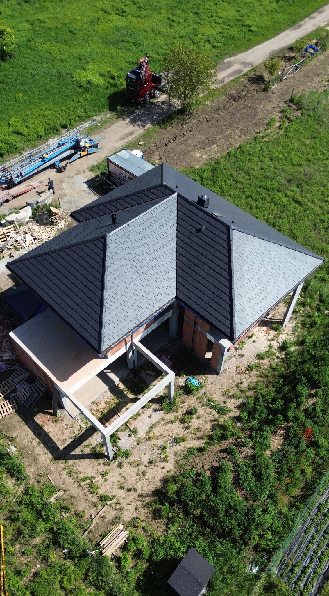 Nowoczesny dom w budowie z ciemnoszarą dachówką, widziany z góry. Na działce widoczny dźwig i materiały budowlane, a w tle rozległa zieleń.