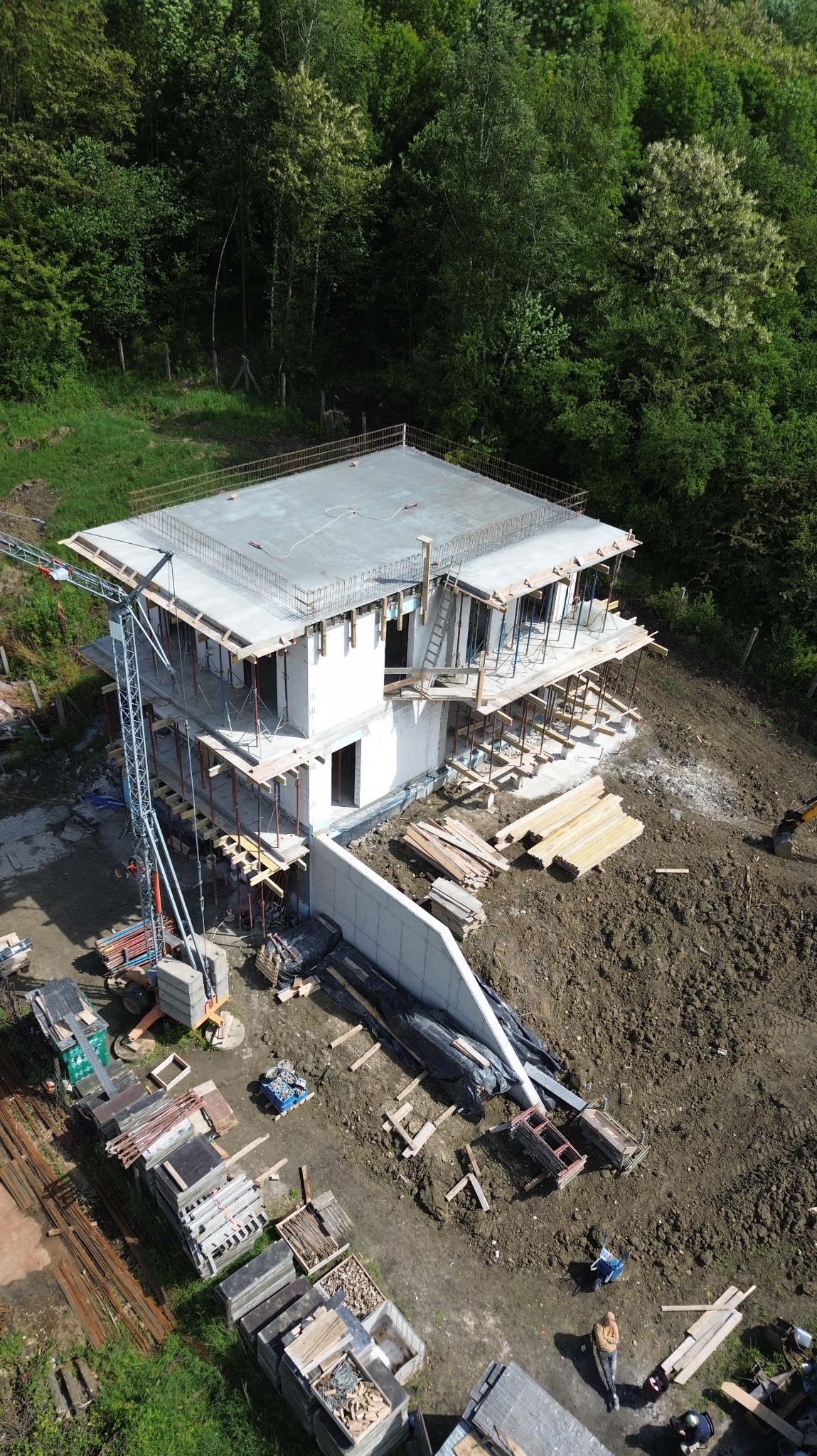 Wysoka perspektywa na budowę domu w stanie surowym otwartym z żurawiem, materiałami budowlanymi i częściowo wylanym fundamentem. Otoczenie to drzewa i ziemia.
