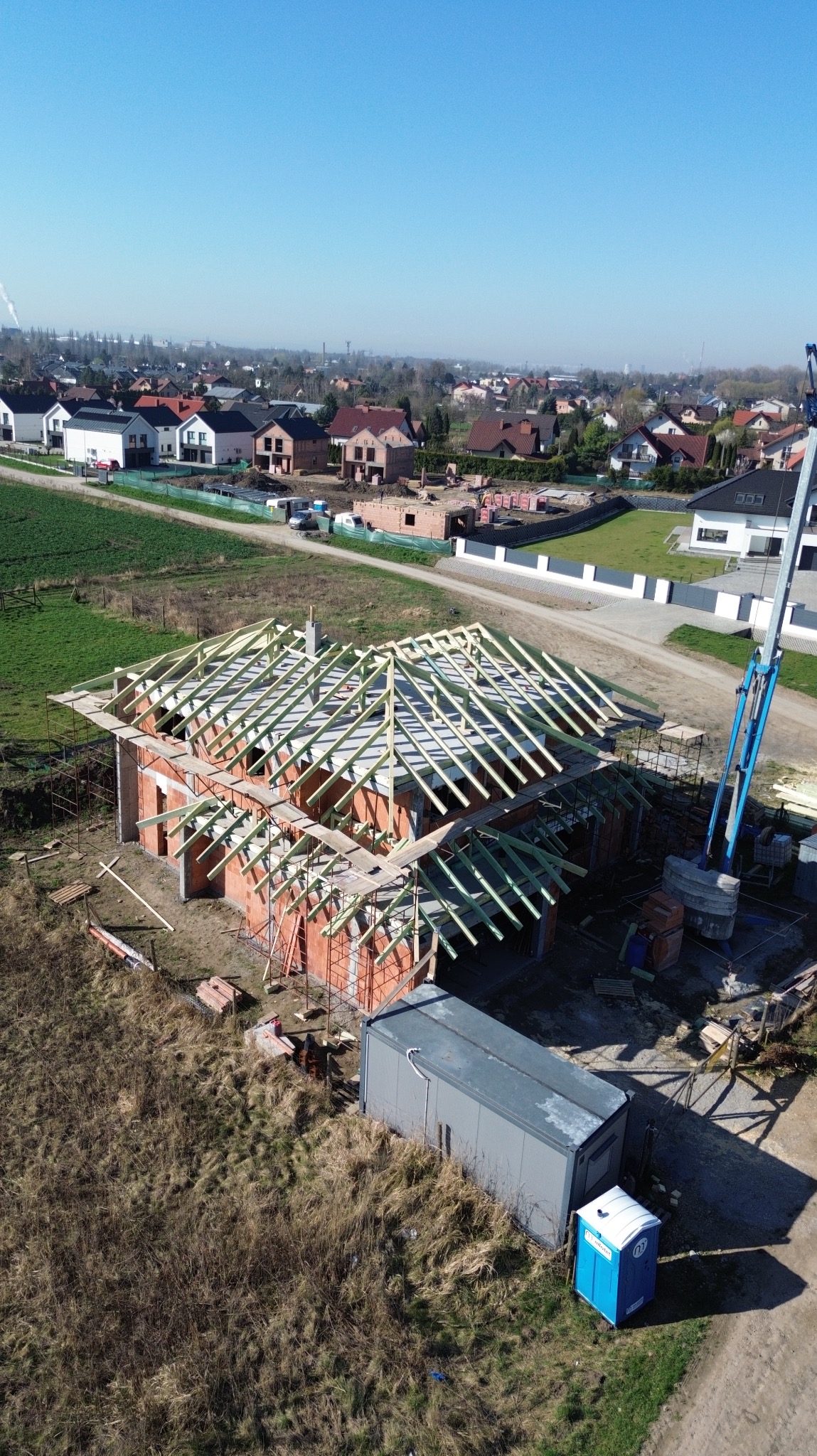 Wysoka perspektywa na budowę domu z czerwonej cegły z drewnianą konstrukcją dachu w trakcie realizacji. W tle zabudowa jednorodzinna i błękitne niebo.
