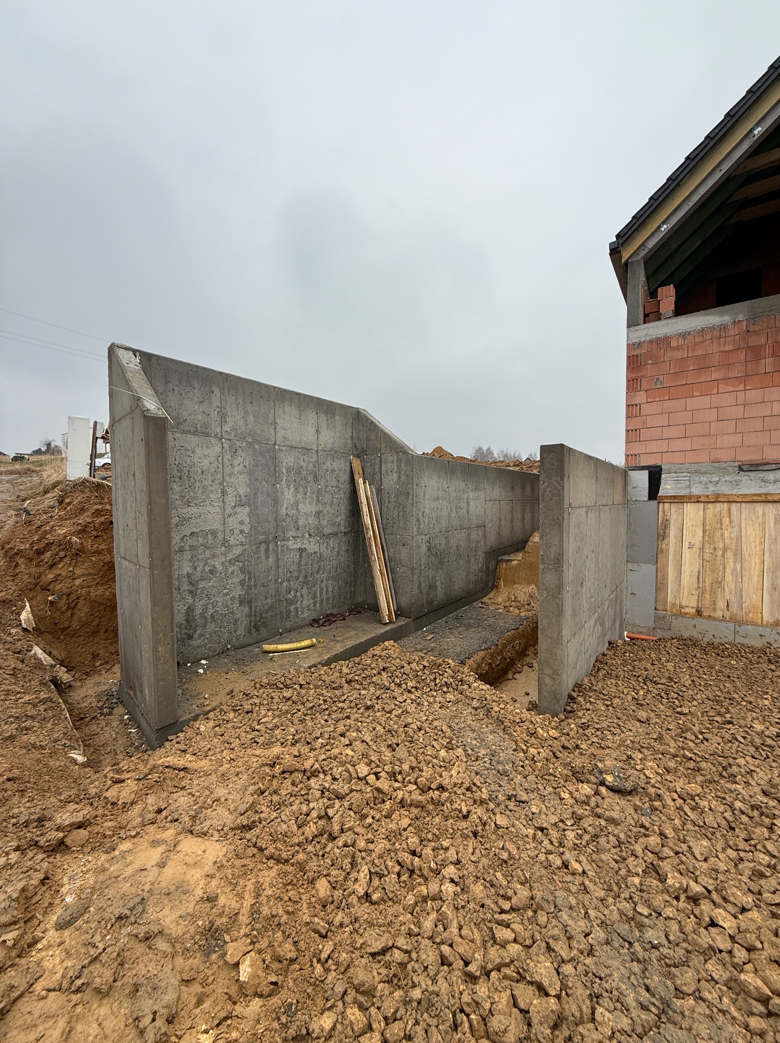 Wykop pod budynek z wylanymi betonowymi fundamentami. Ściany fundamentowe z betonu, widoczne zbrojenia. Część budynku w trakcie budowy, mur z czerwonej cegły.