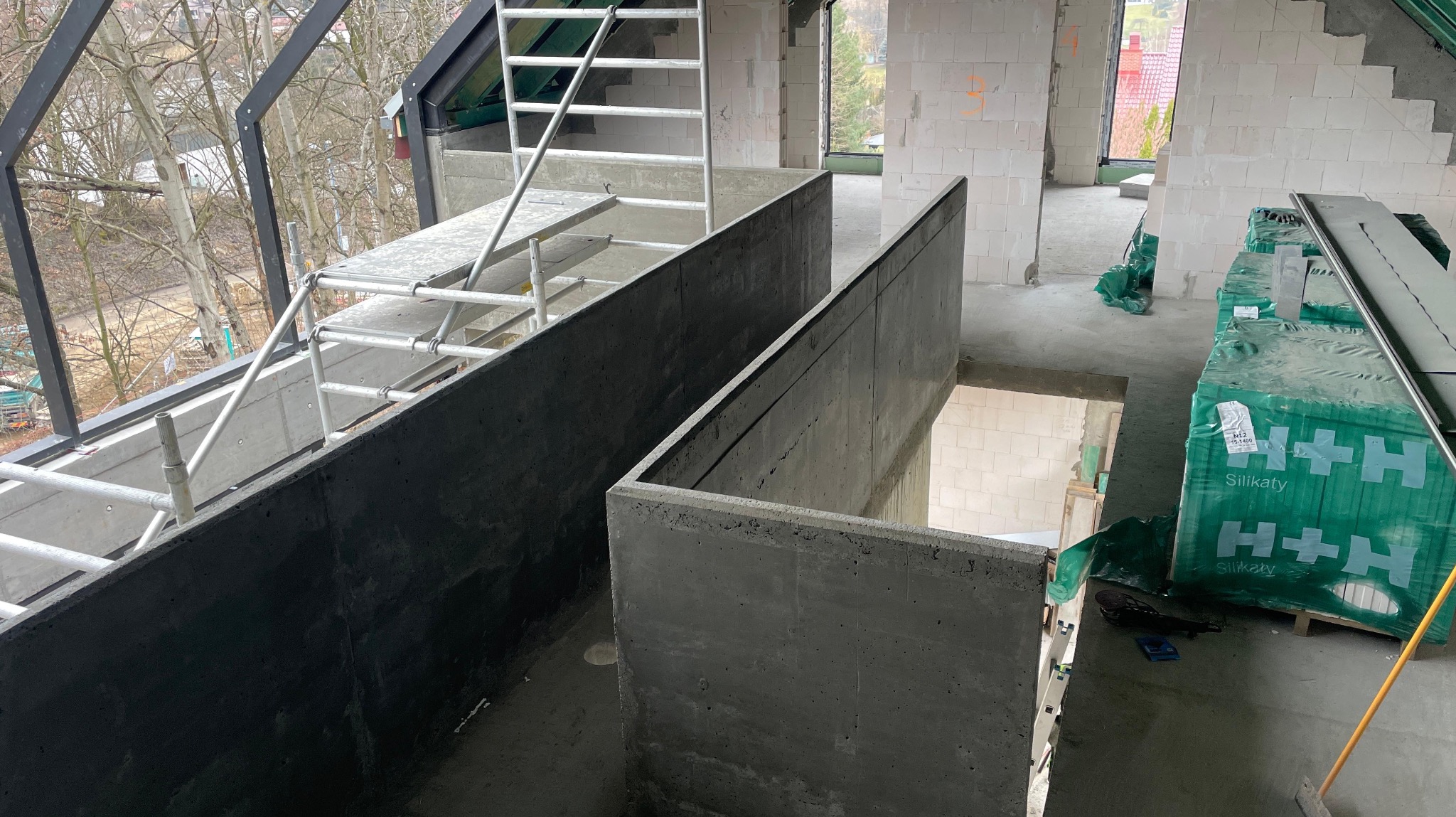 Wnętrze domu w trakcie budowy: surowe ściany z betonu komórkowego, rusztowanie, palety z bloczkami H+H. Widok z góry na betonowe elementy konstrukcyjne.