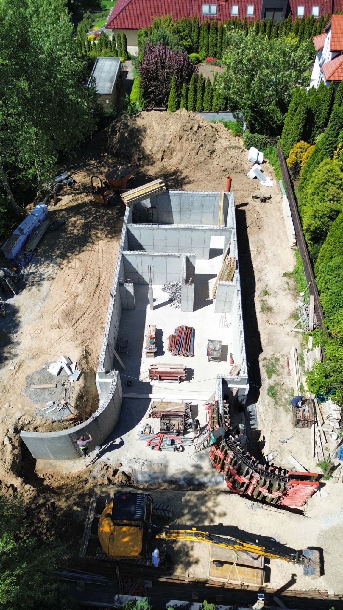 Fundamenty domu z betonu, widok z góry. Ustawione szalunki do wylania łukowatej ściany. Żółta koparka na placu budowy, w tle domy jednorodzinne i zieleń.