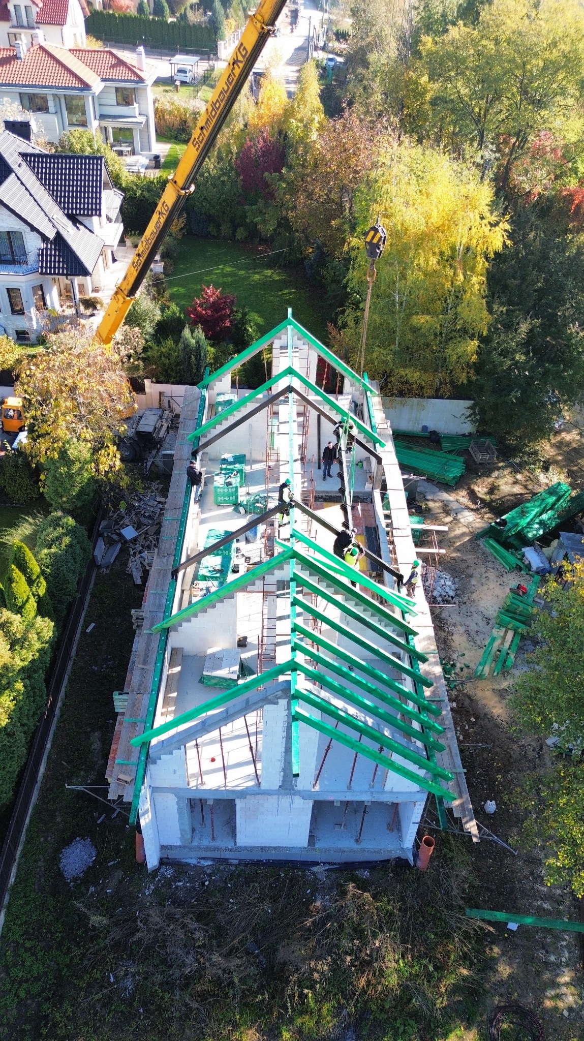 Budowa domu z białych bloczków: widok z góry na wznoszone ściany i konstrukcję dachu z zielonymi elementami. Żuraw budowlany w tle. Jesienna sceneria.