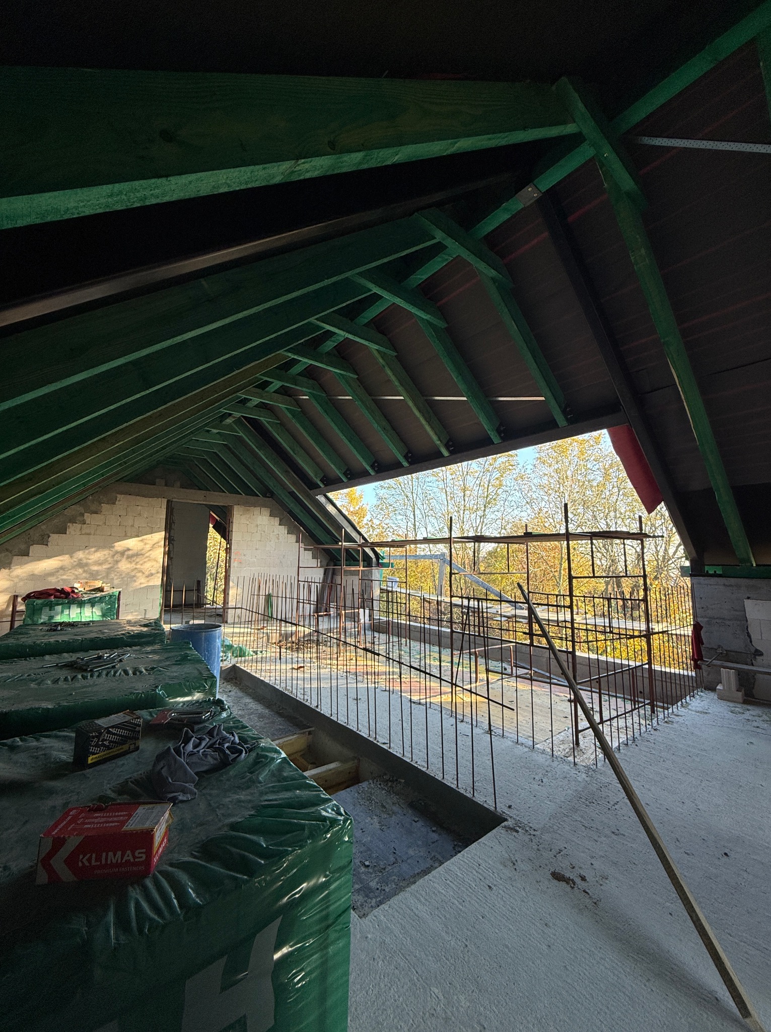 Wnętrze poddasza w trakcie budowy: zielona więźba dachowa, bloczki, zbrojenie, widok na jesienne drzewa przez otwór w ścianie. Elementy mocujące Klimas na pierwszym planie.