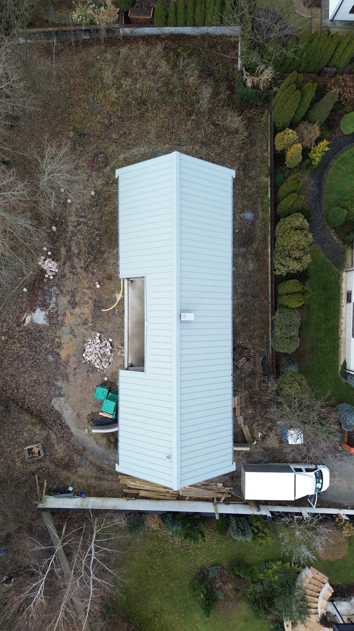 Nietypowy widok z drona na dom w trakcie budowy z szarą elewacją i fragmentem dachu. Widoczny niezabudowany otwór okienny i biały samochód dostawczy.