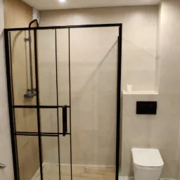 Nowoczesna kabina prysznicowa z czarną ramą i szklanymi drzwiami, widoczny fragment toalety podwieszanej oraz terazzo na podłodze, jasne płytki na ścianach.