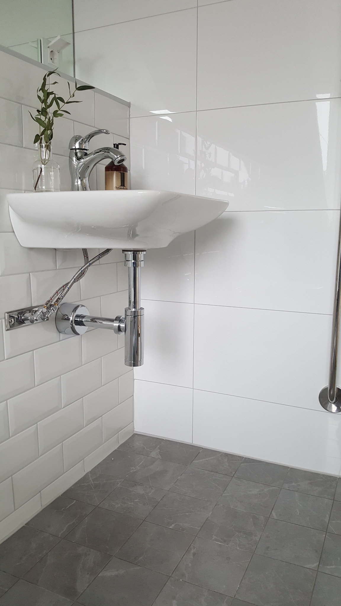 Nowoczesna łazienka z białą umywalką, armaturą chromowaną i szarymi płytkami podłogowymi imitującymi marmur, uzupełniona o minimalistyczny flakon z zielonymi gałązkami.