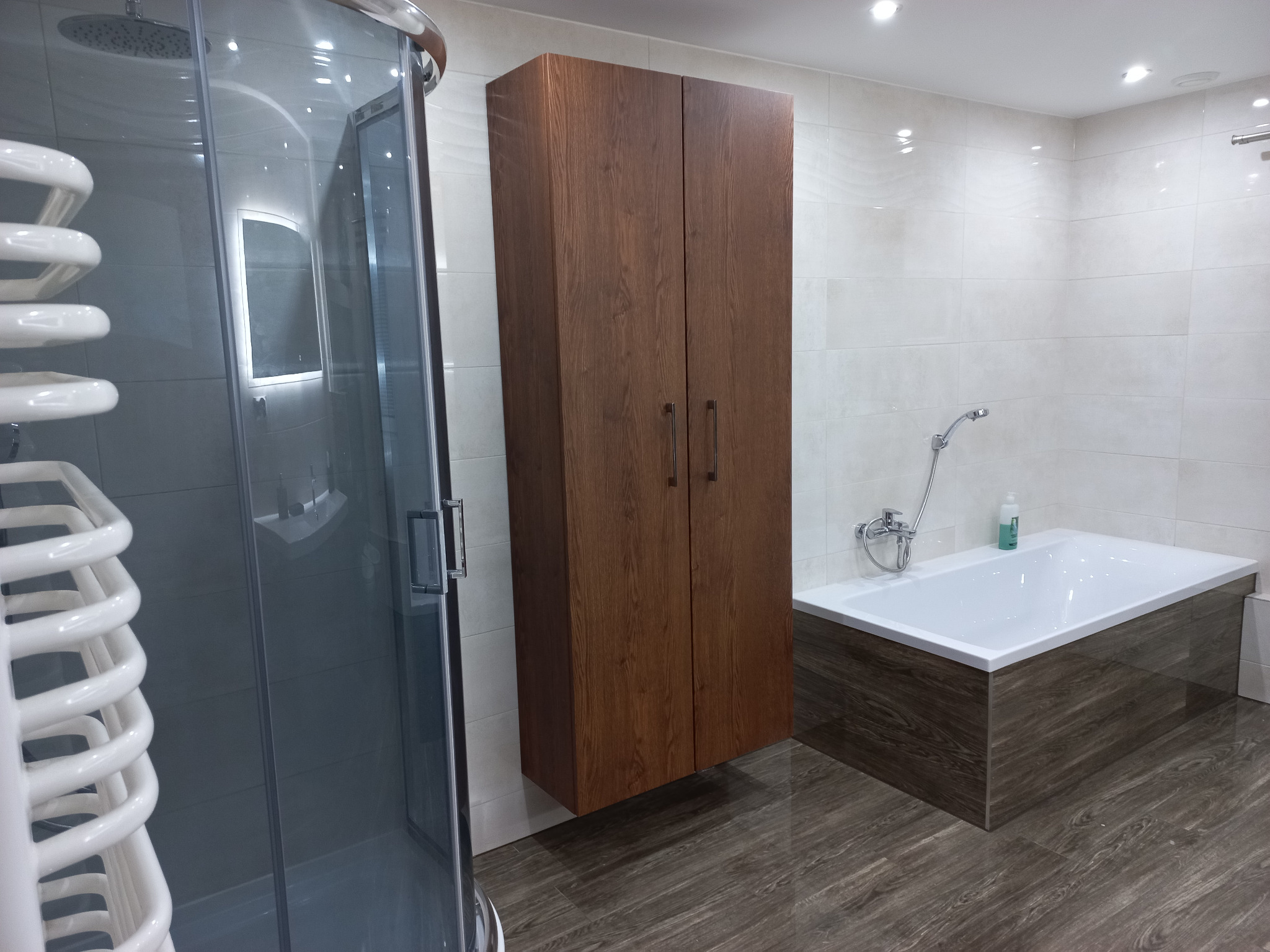 Nowoczesna łazienka z kabiną prysznicową, wiszącą szafką z ciemnego drewna i wanną obudowaną panelami w odcieniach brązu, podłoga w kolorze ciemnego drewna.