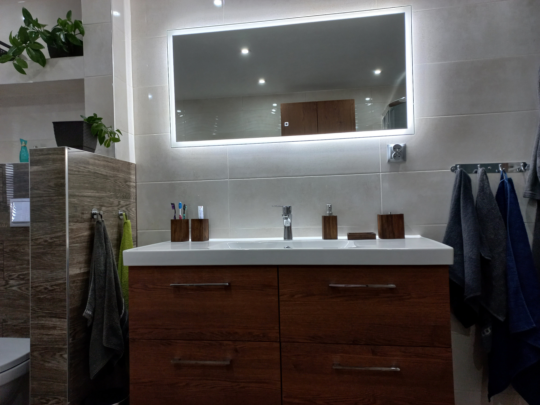 Nowoczesna łazienka z drewnianą szafką pod umywalką, podświetlanym lustrem i szarymi płytkami na ścianach, akcent roślinny w postaci doniczek na półce.