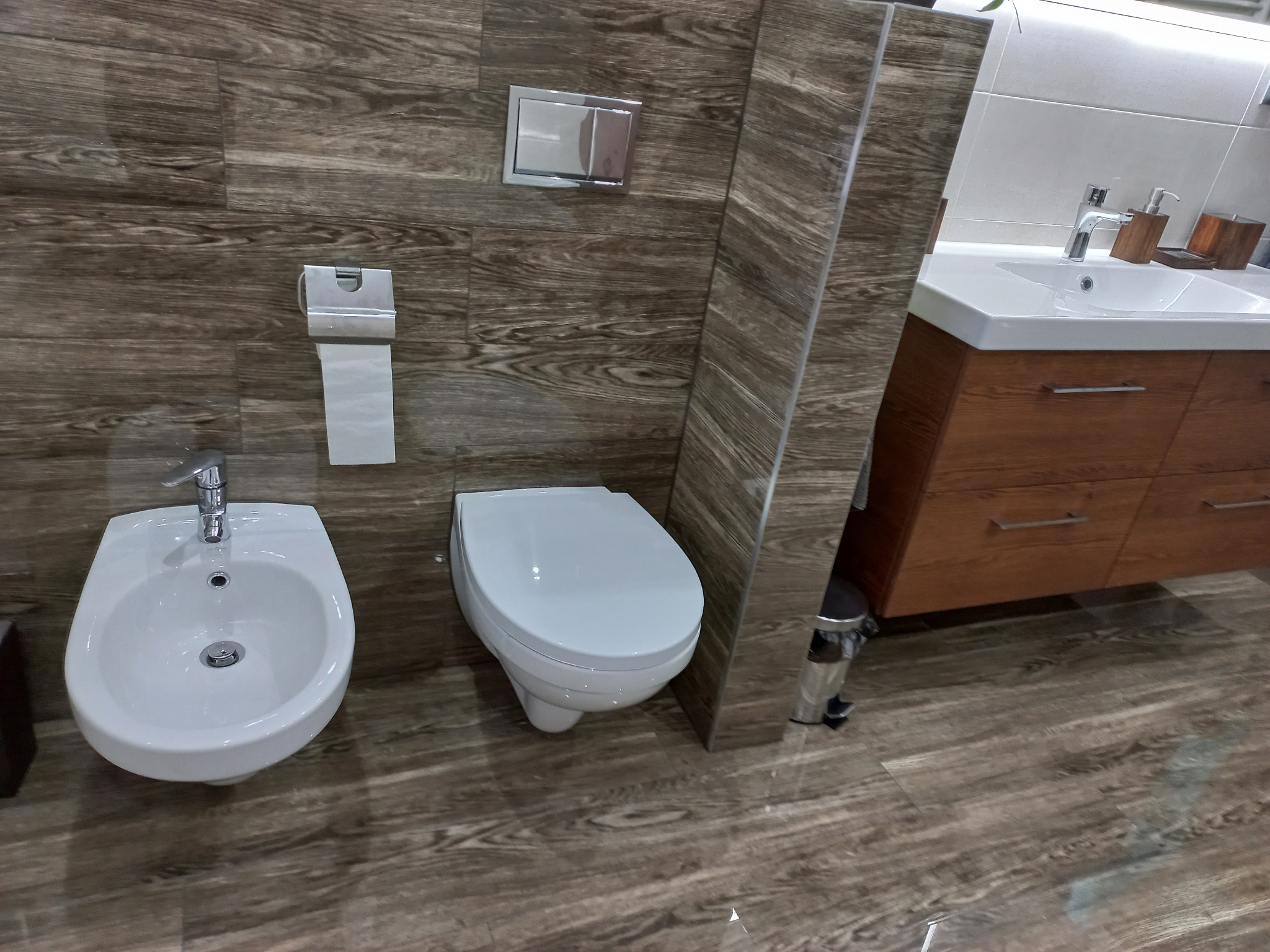 Nowoczesna łazienka z drewnopodobnymi płytkami, wiszącą miską WC, bidetem i szafką pod umywalką z drewnianymi frontami.