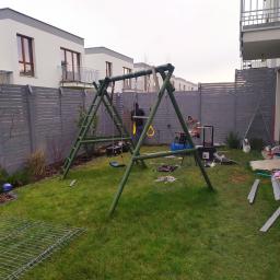 PolixBud - Montaż placu zabaw: drewniana huśtawka w trakcie instalacji na trawniku przed nowym domem z szarym ogrodzeniem w tle, widoczne narzędzia i elementy konstrukcyjne.