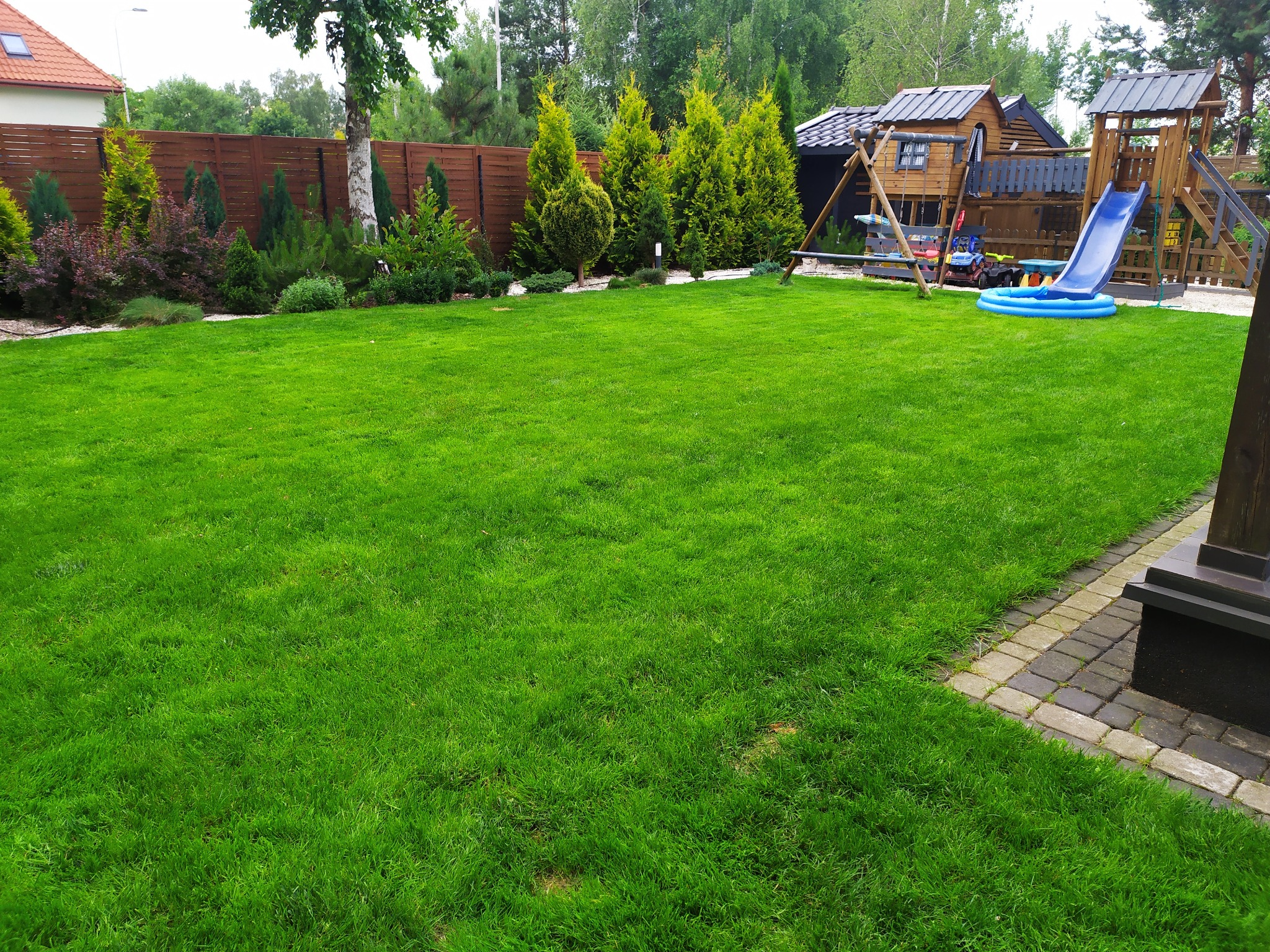 Zadbany trawnik z placem zabaw dla dzieci, otoczony tujami i drewnianym ogrodzeniem, z widocznym fragmentem kostki brukowej.