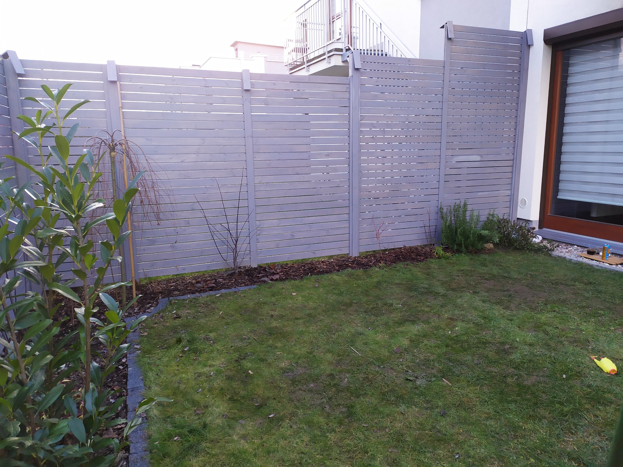 Szare, poziome ogrodzenie panelowe z drewnianych listew, oddzielające trawnik od posesji sąsiada, z widocznym fragmentem domu z balkonem i oknem z roletami.