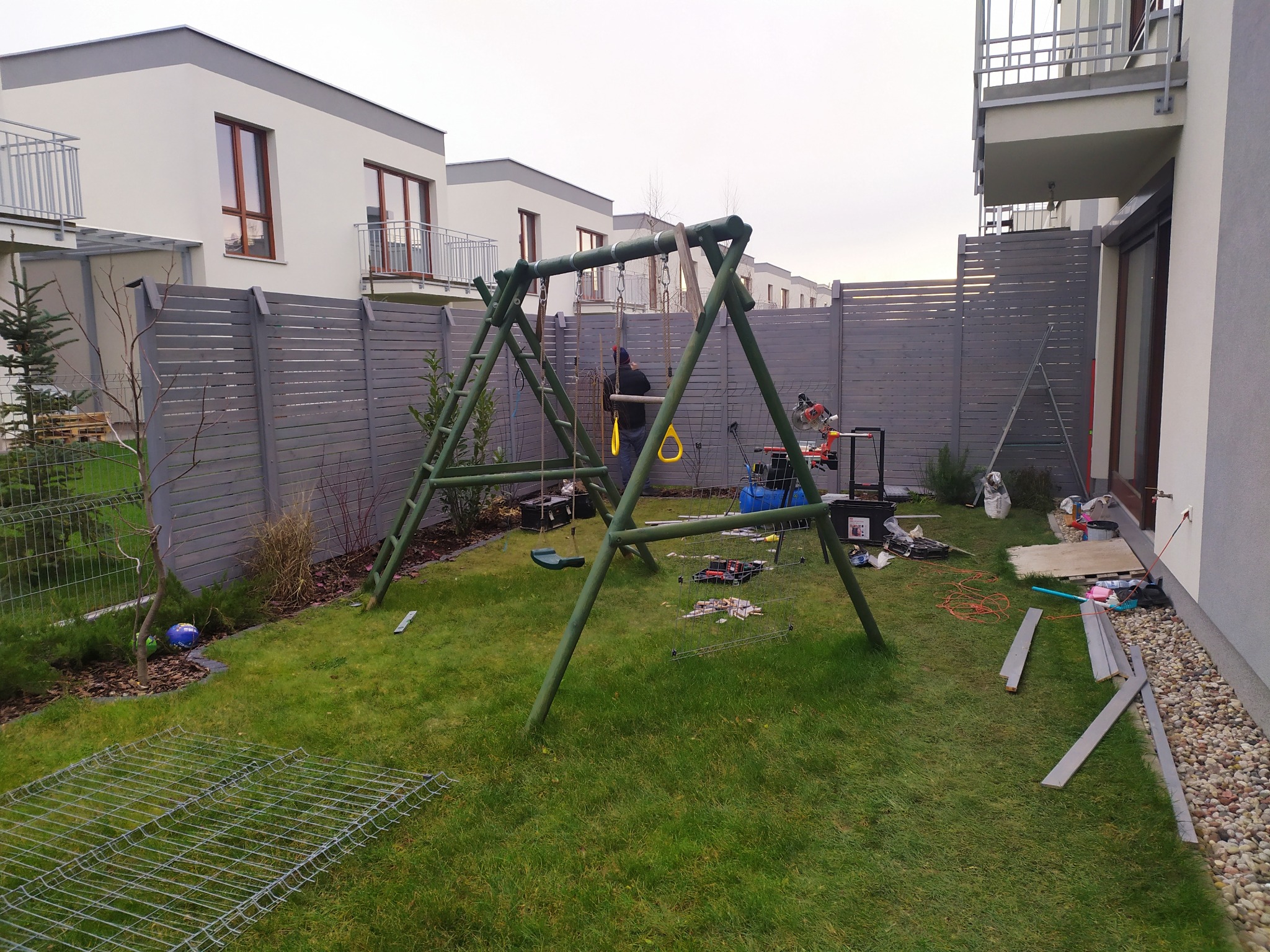 Montaż placu zabaw: drewniana huśtawka w trakcie instalacji na trawniku przed nowym domem z szarym ogrodzeniem w tle, widoczne narzędzia i elementy konstrukcyjne.