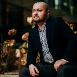 Portret mężczyzny w marynarce i kraciastej koszuli, siedzącego na tle nowoczesnego budynku i uschniętych kwiatów, spoglądającego w lewo.