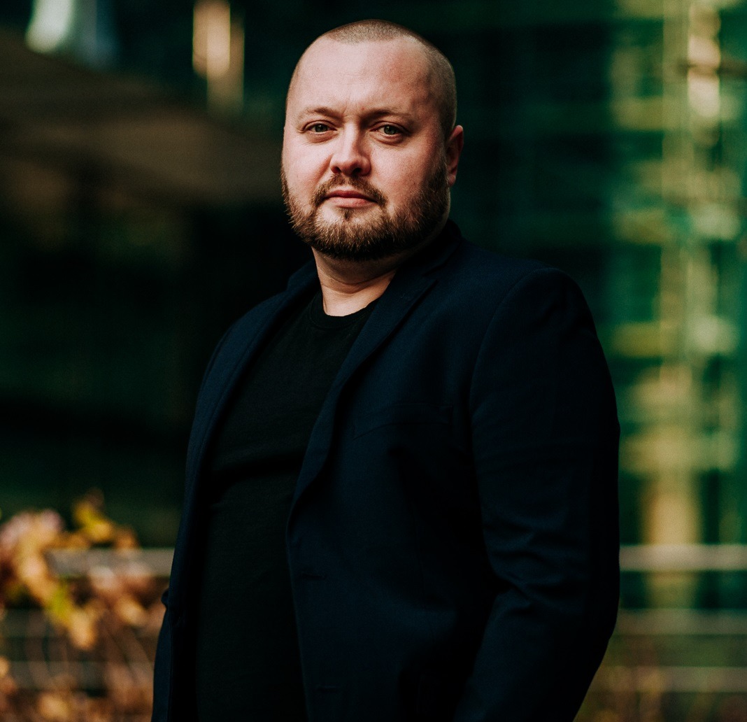 Portret mężczyzny w ciemnej marynarce na tle zielonej fasady budynku.