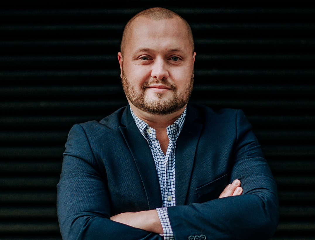 Portret mężczyzny w granatowej marynarce i wzorzystej koszuli, z założonymi rękami na tle czarnej, poziomej struktury.